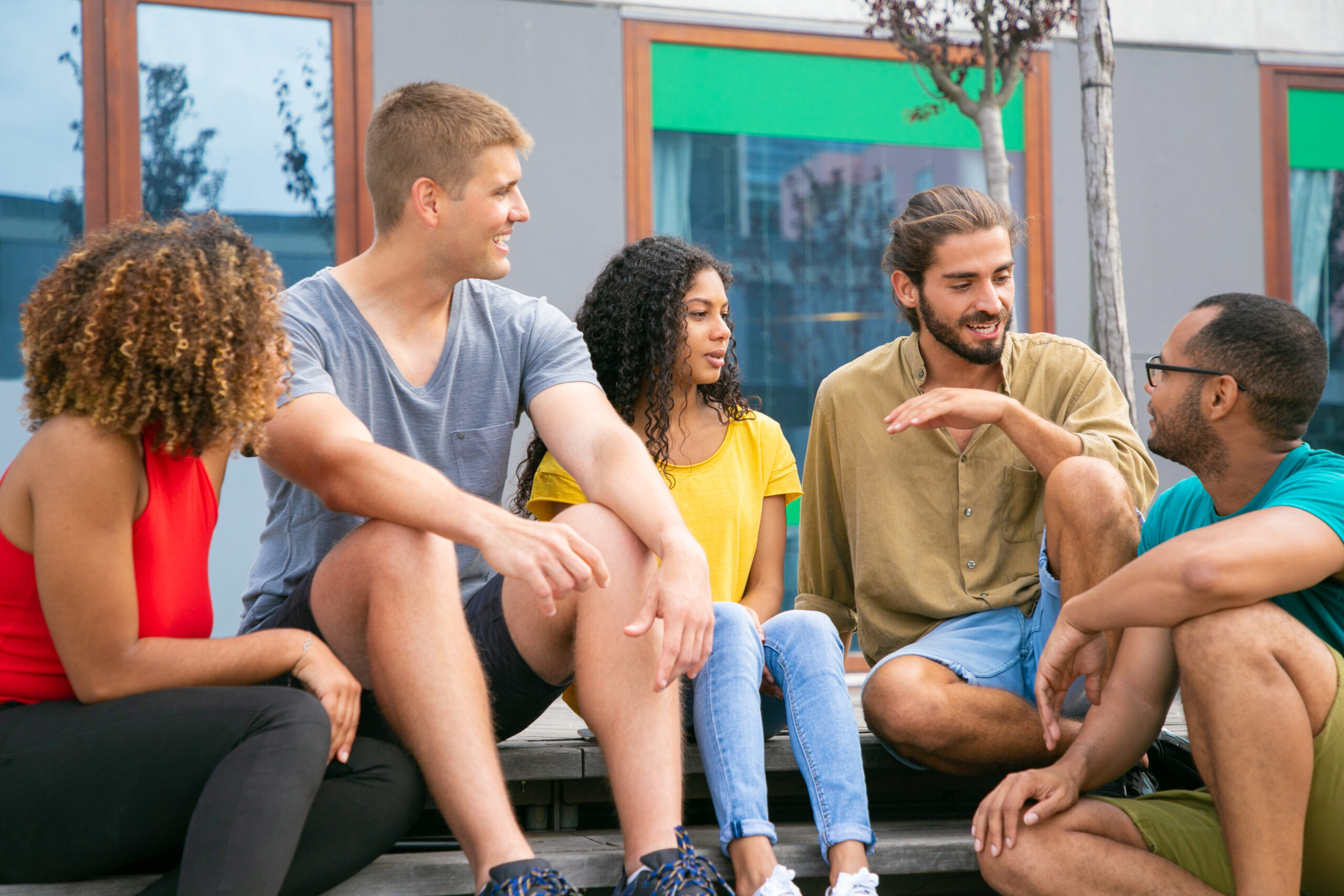 Content young friends sitting on steps. Cheerful young multiethnic men and women sitting together on street and interacting. Friendship concept
