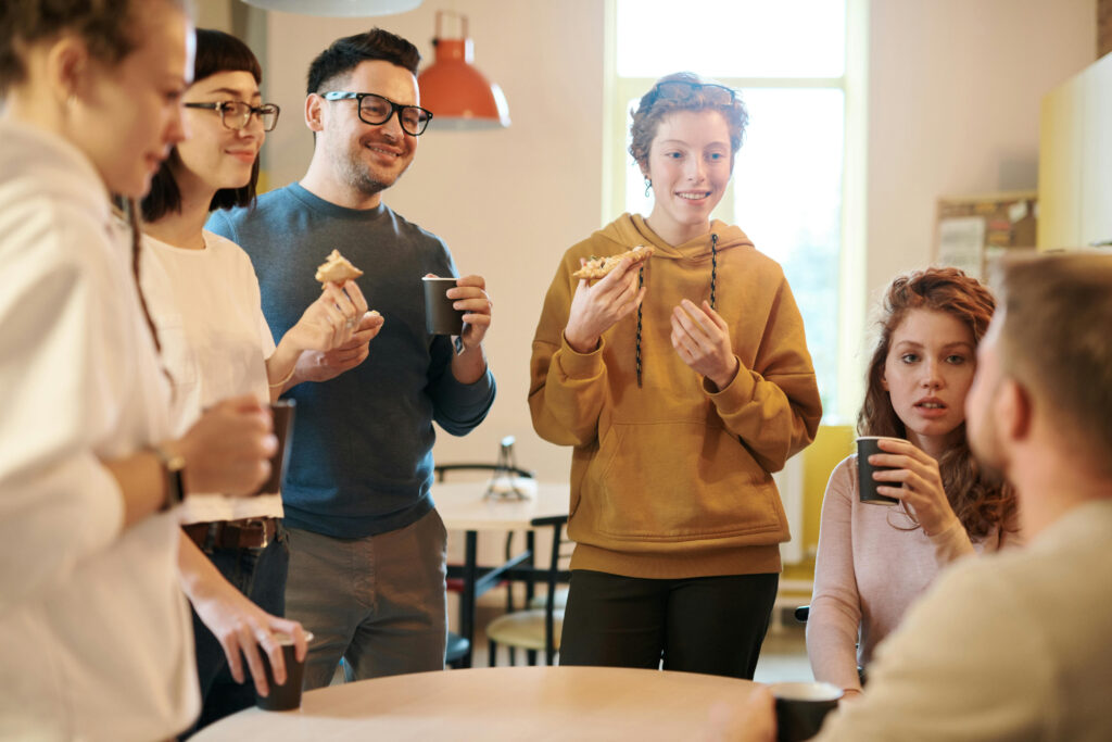 Mehrere Menschen stehen um einen Tisch, essen etwas und sprechen miteinander